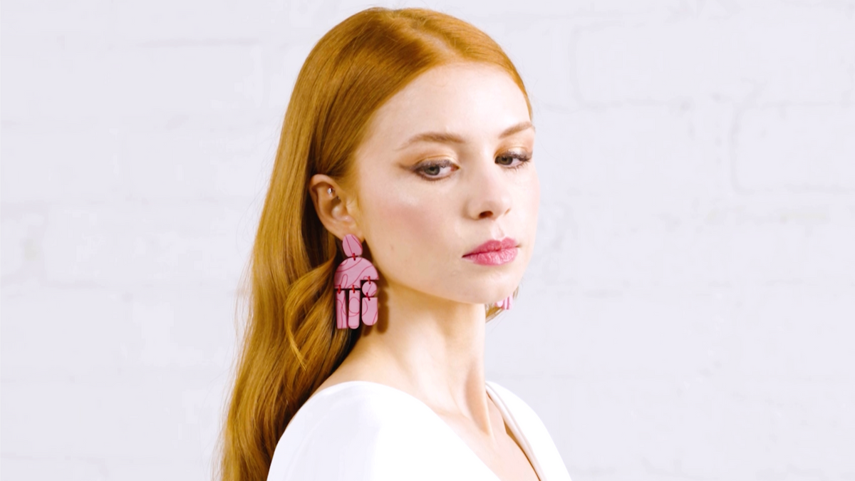 A woman with long red hair wearing a white top and pink earrings, photographed against a white wall.