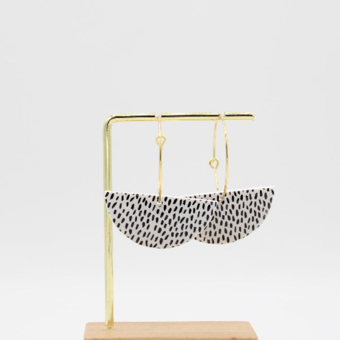 A pair of crescent-shaped dangle earrings with a black and white spotted pattern, hanging from gold-tone rings on a gold display stand with a wooden base against a white background.