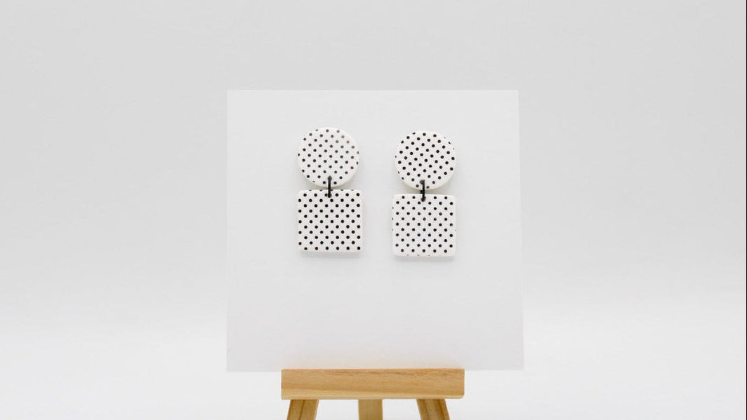 A pair of black and white polka dot earrings displayed on a white card mounted on a small wooden easel against a light gray background.