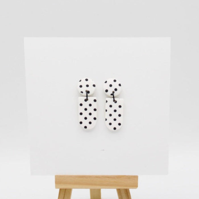 A pair of white and black polka dot earrings displayed on a white card mounted on a wooden stand against a light gray background.