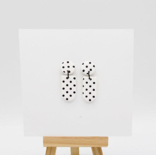 A pair of white and black polka dot earrings displayed on a white card mounted on a wooden stand against a light gray background.