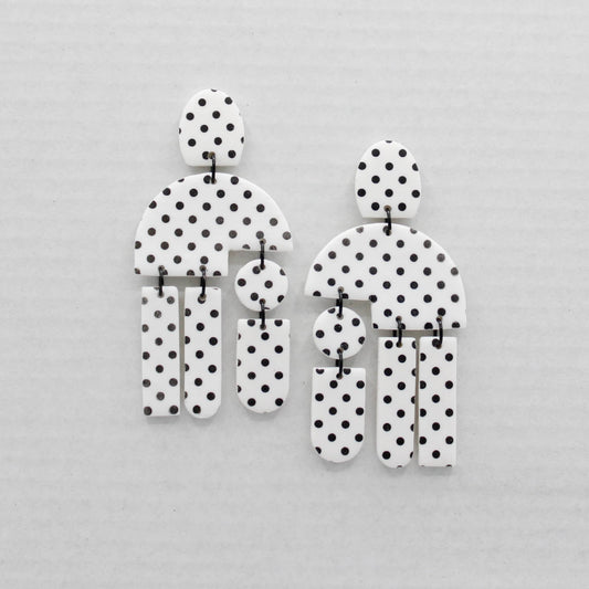 Two geometric shape earrings with a black and white polka dot pattern, displayed on a light gray textured background.