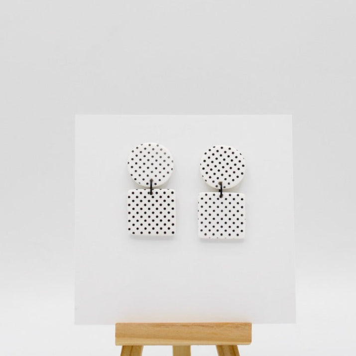 A pair of black and white polka dot earrings displayed on a white card mounted on a small wooden easel against a light gray background.